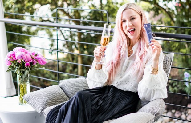 Renee Rouleau sitting with a glass of champagne in one hand and a Renee Rouleau skincare product in the other in a happy mood